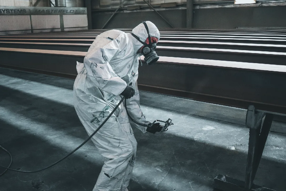 Trabalhador com equipamento de proteção pintando vigas metálicas em uma fábrica. — Medicina Ocupacional Curitiba