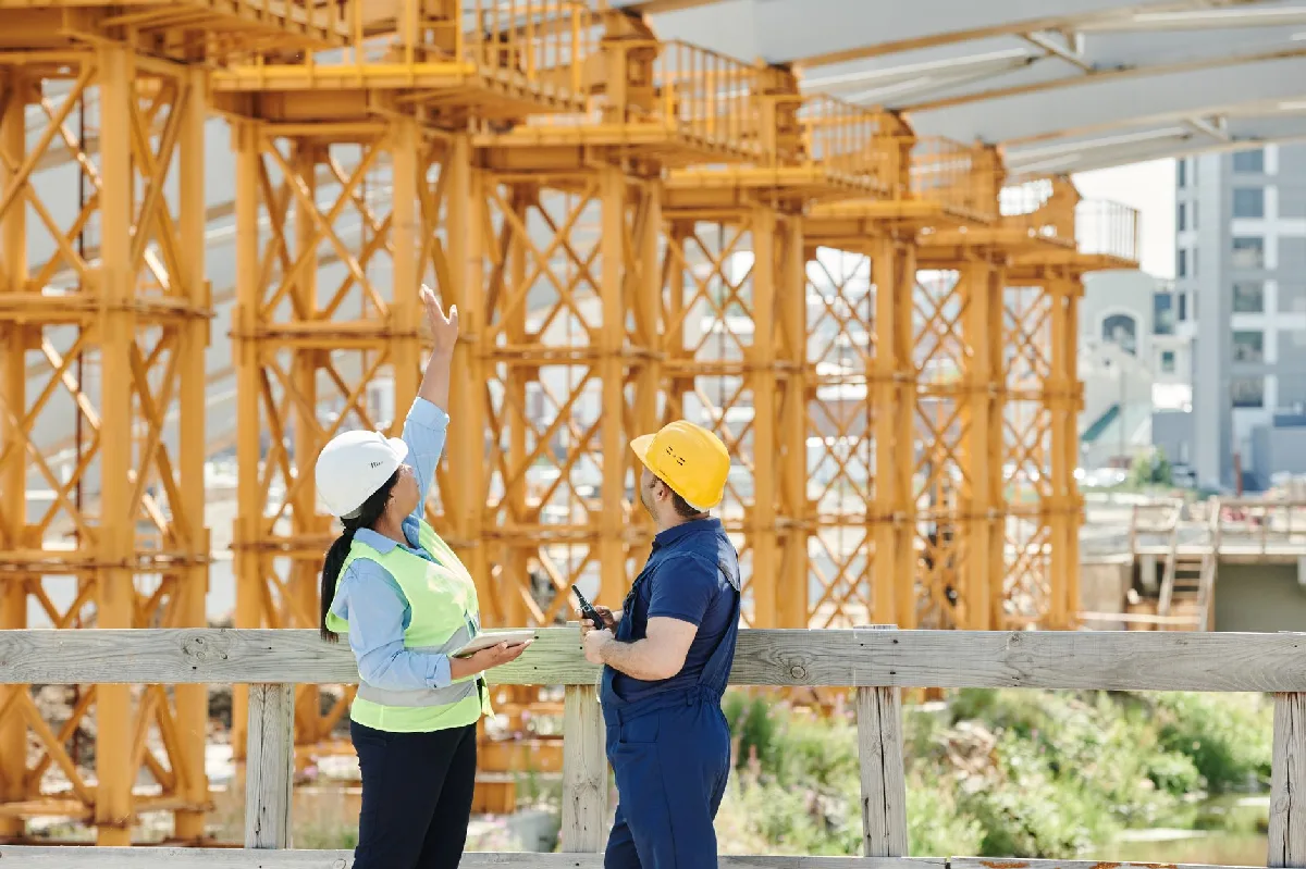 Dois engenheiros de capacete discutem projetos em um canteiro de obras movimentado. — Medicina Ocupacional Curitiba