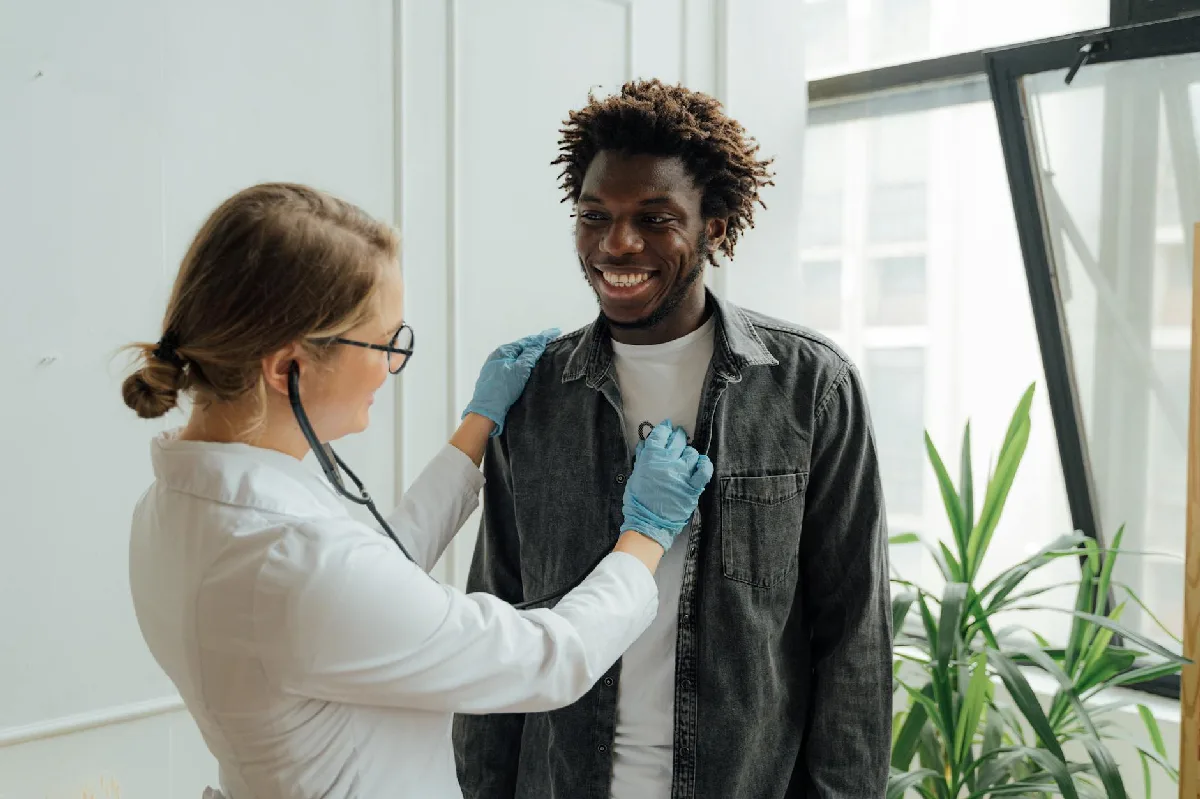 Coordenação de PCMSO: prazos, NRs e responsabilidades em Curitiba Médico com estetoscópio examina paciente sorrindo em ambiente interno, promovendo saúde e cuidado. — Medicina Ocupacional Curitiba
