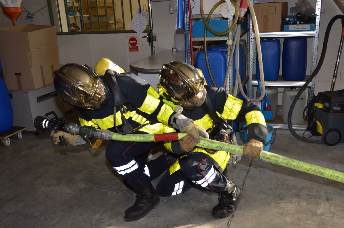 Ficha de EPI para indústrias de Curitiba: Gestão Legal Direta Dois bombeiros em equipamento completo manuseiam uma mangueira de incêndio dentro de um armazém. — Medicina Ocupacional Curitiba