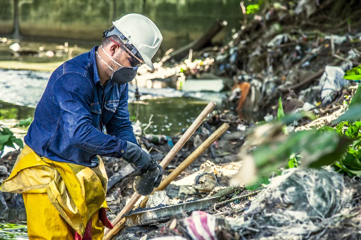 Trabalhador de segurança limpa margens de rio poluído, focado em proteção ambiental. — Medicina Ocupacional Curitiba