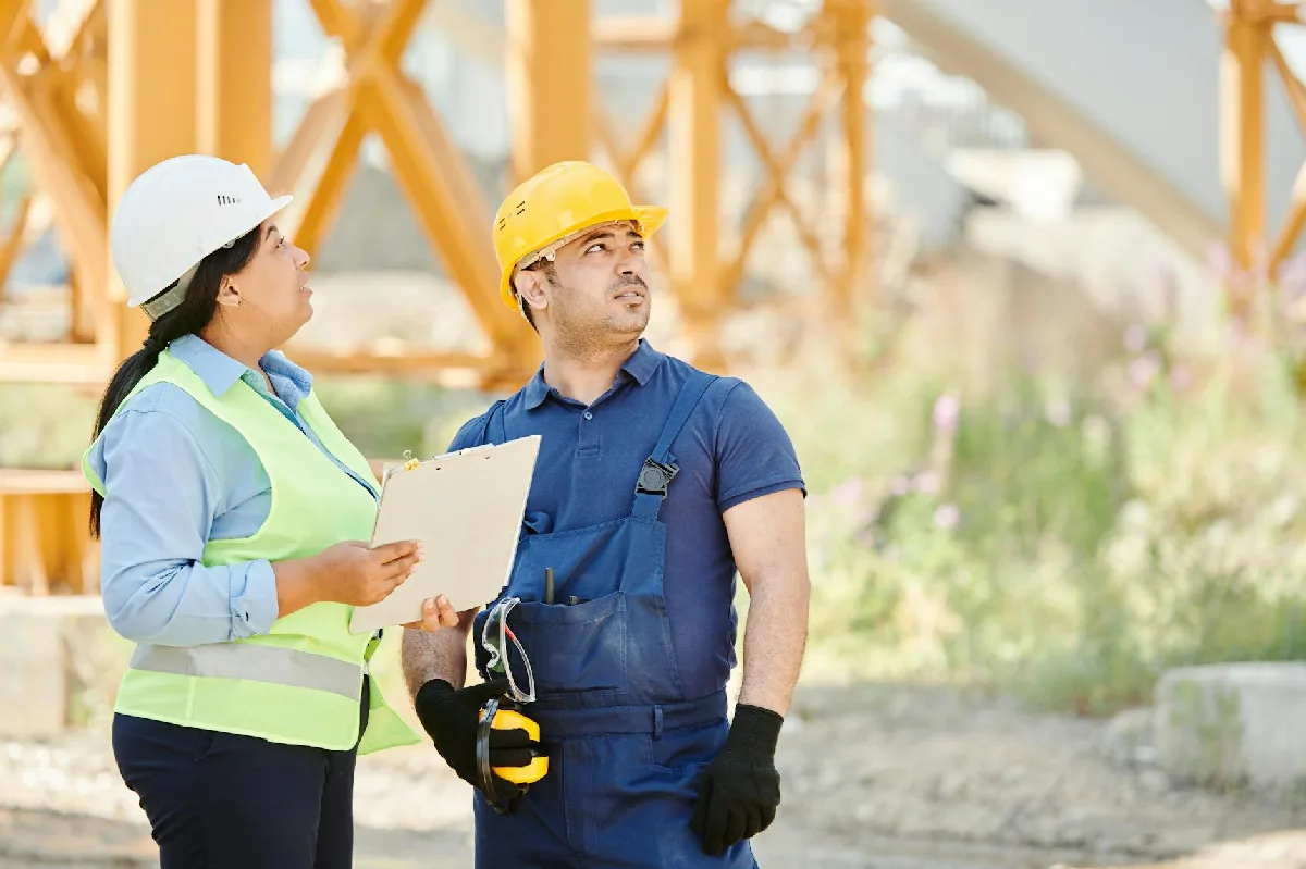 Dois trabalhadores da construção civil com equipamentos de segurança discutem planos em um canteiro de obras. — Medicina Ocupacional Curitiba