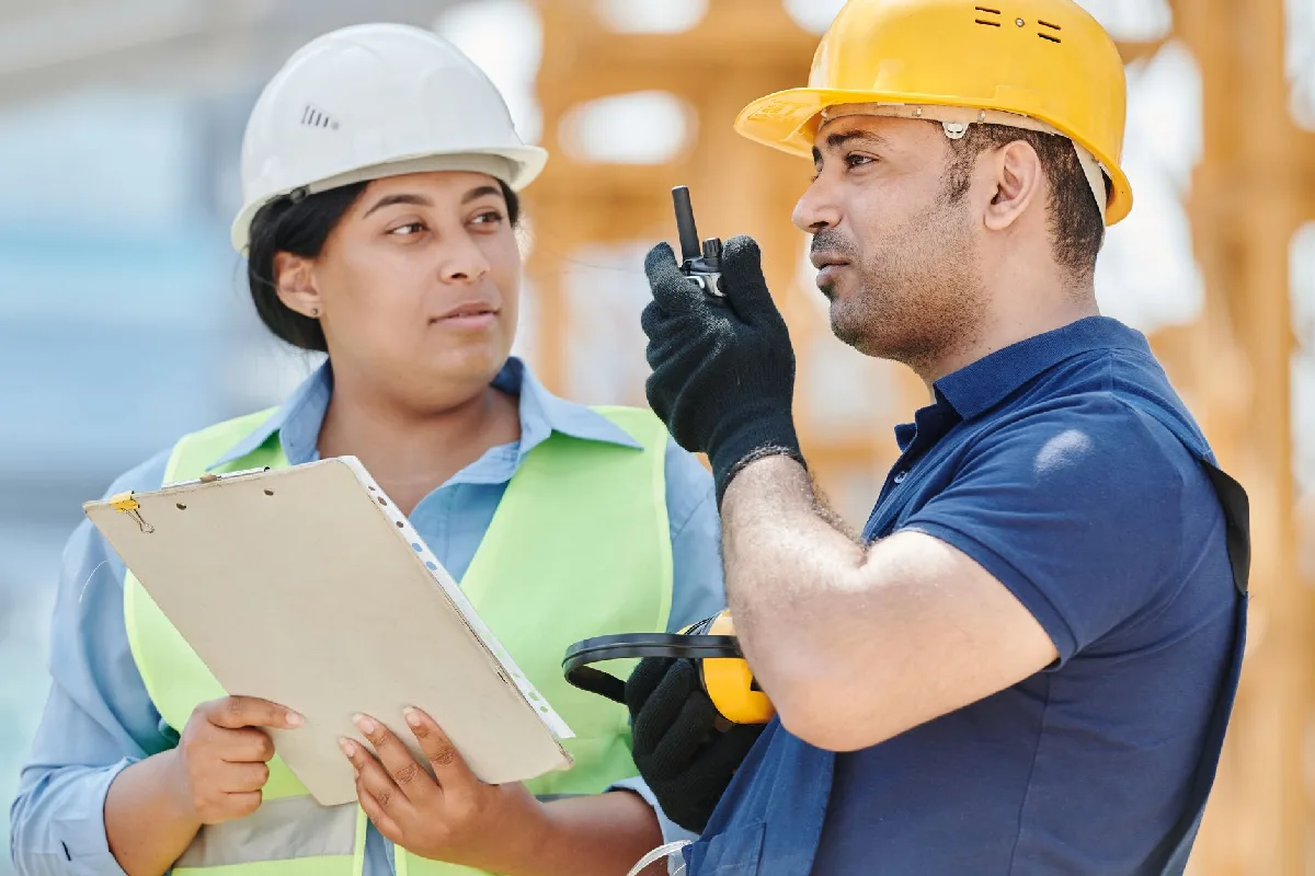 Plano de Ação NR-01 Curitiba: o que sua empresa precisa cumprir Dois trabalhadores da construção civil com equipamentos de segurança discutem planos em um canteiro de obras. — Medicina Ocupacional Curitiba