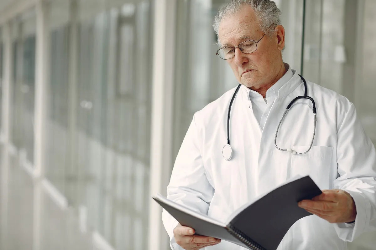 Programa de Controle Médico: prazos, NRs e responsabilidades em Curitiba Médico em jaleco branco com estetoscópio lê prontuários em corredor de hospital moderno. — Medicina Ocupacional Curitiba